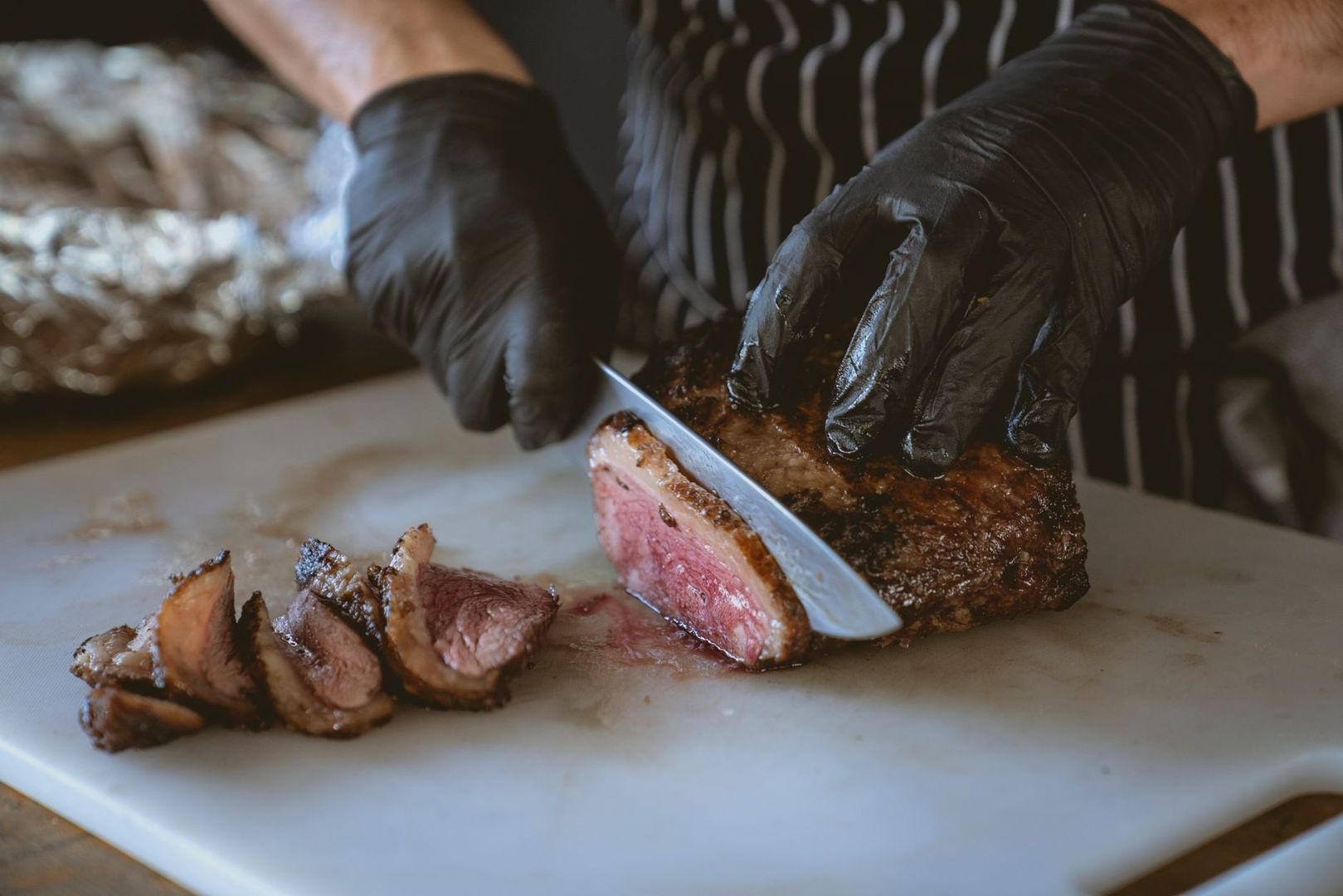 Kock i svarta handskar skär en köttbit på skärbräda